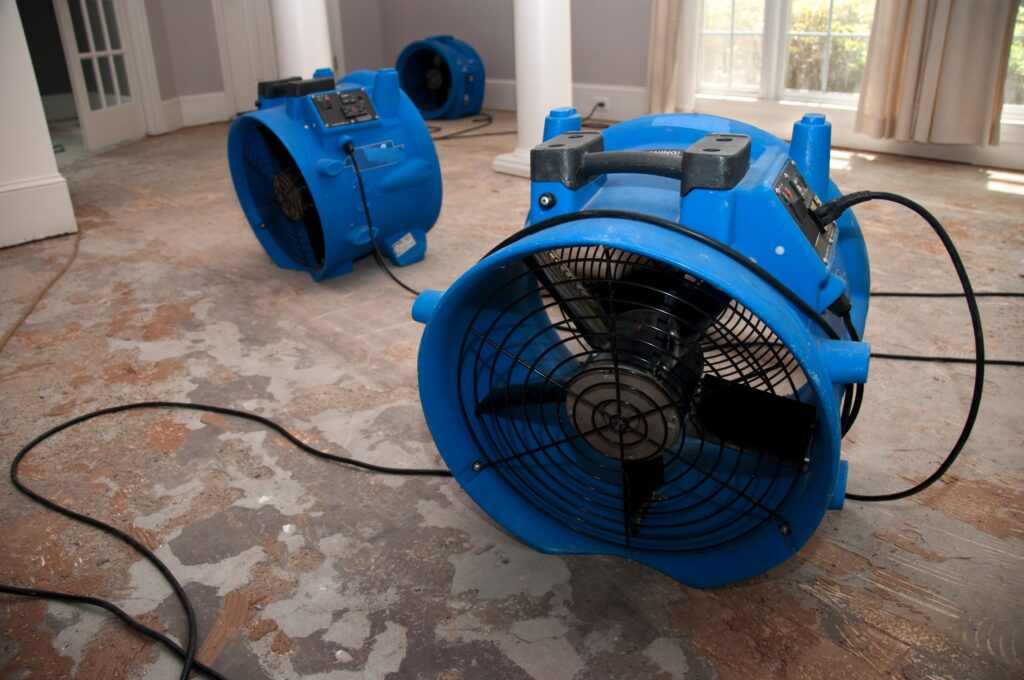 Industrial fans and dehumidifiers in an empty water-damaged room in a home. The flooring is ripped off, and the rooms are sprayed with biowash. 